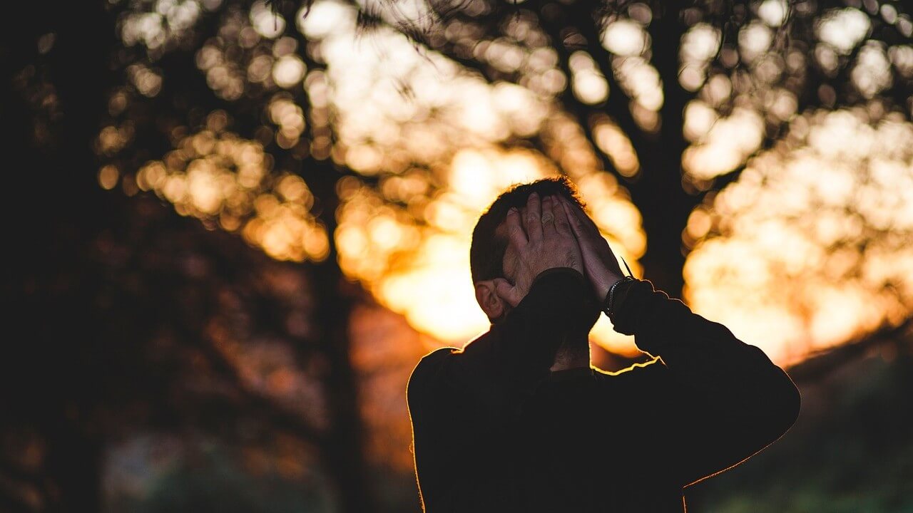 A man sitting with his hands over his face, in turmoil, with the sun settings behind him